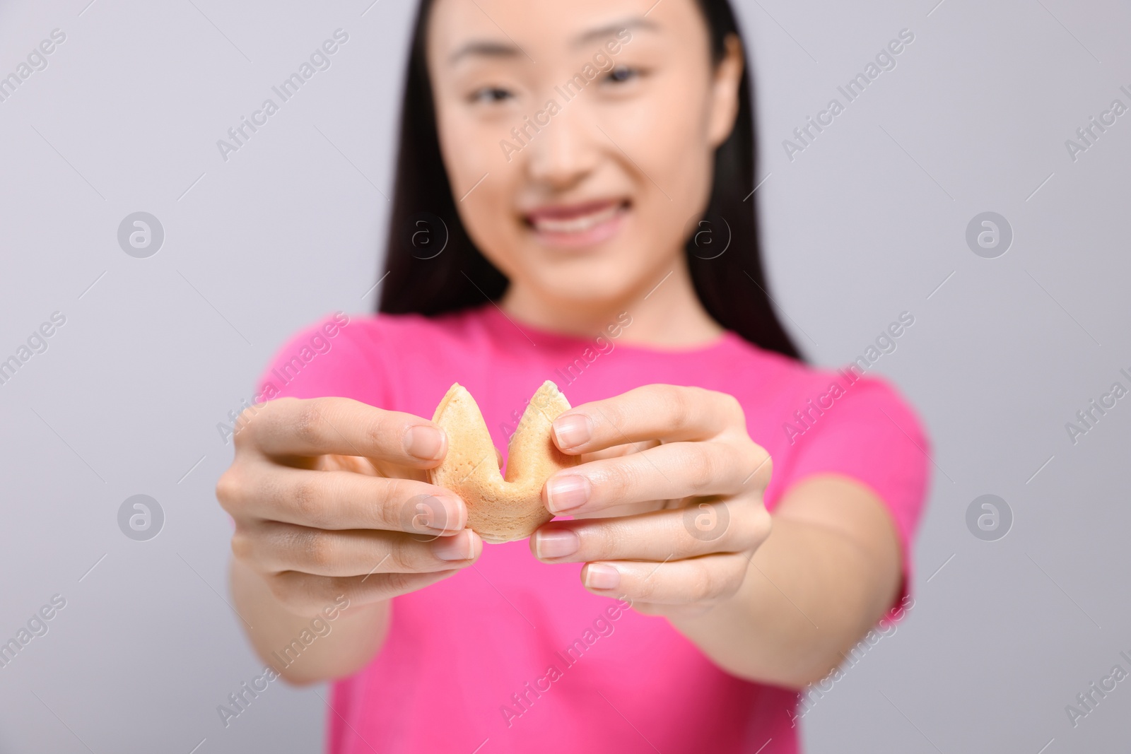 Photo of Asian woman against grey background, focus on tasty fortune cookie with prediction