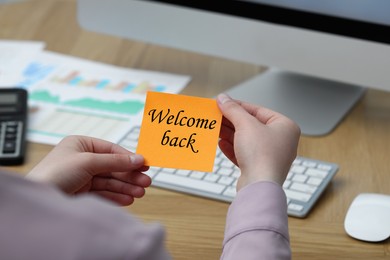 Image of Woman holding paper note with phrase Welcome Back at office desk, closeup