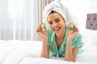 Photo of Young woman with cleansing mask on her face holding cucumber slices in bedroom