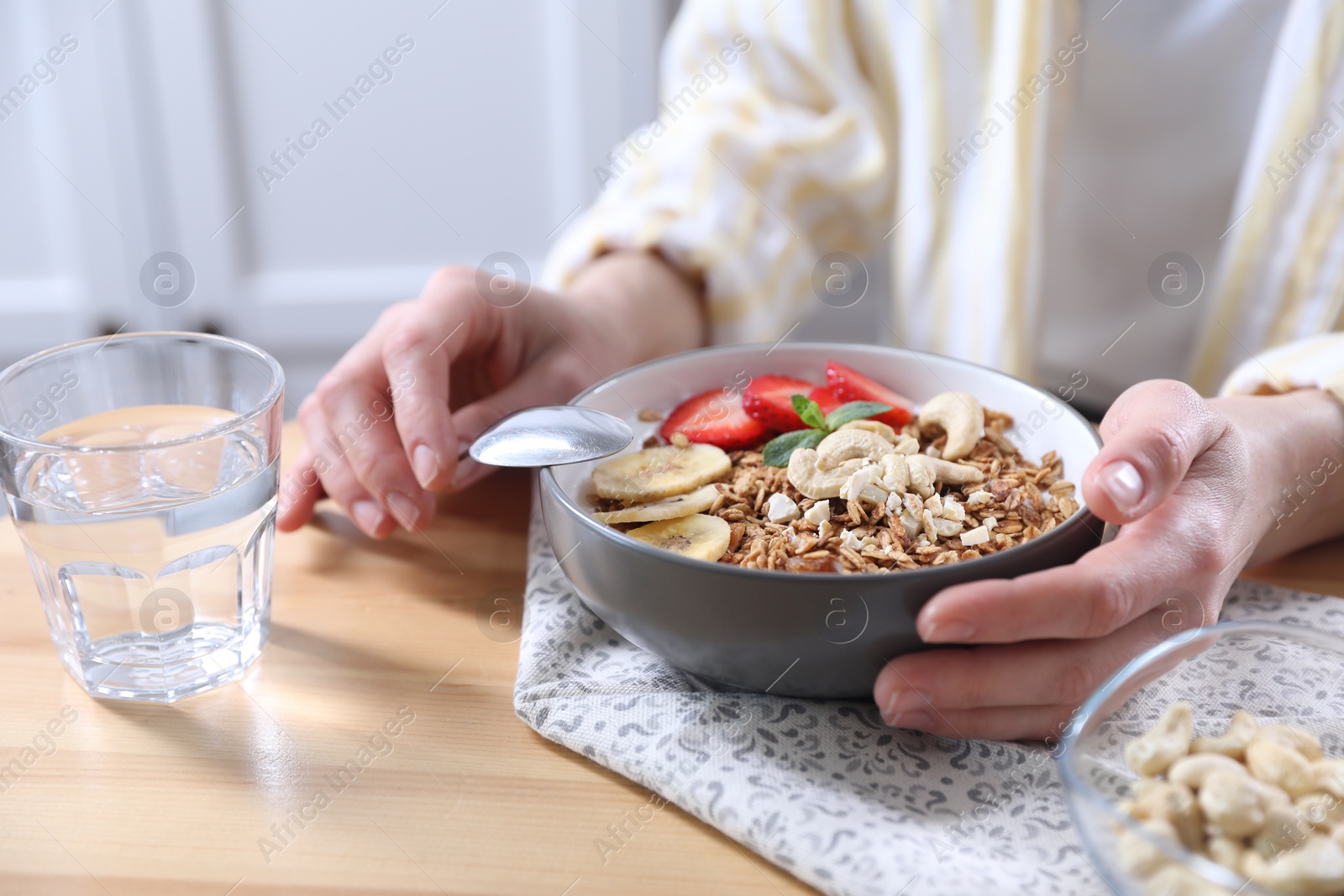 Photo of Woman eating tasty granola with banana, cashew and strawberries at wooden table indoors, closeup