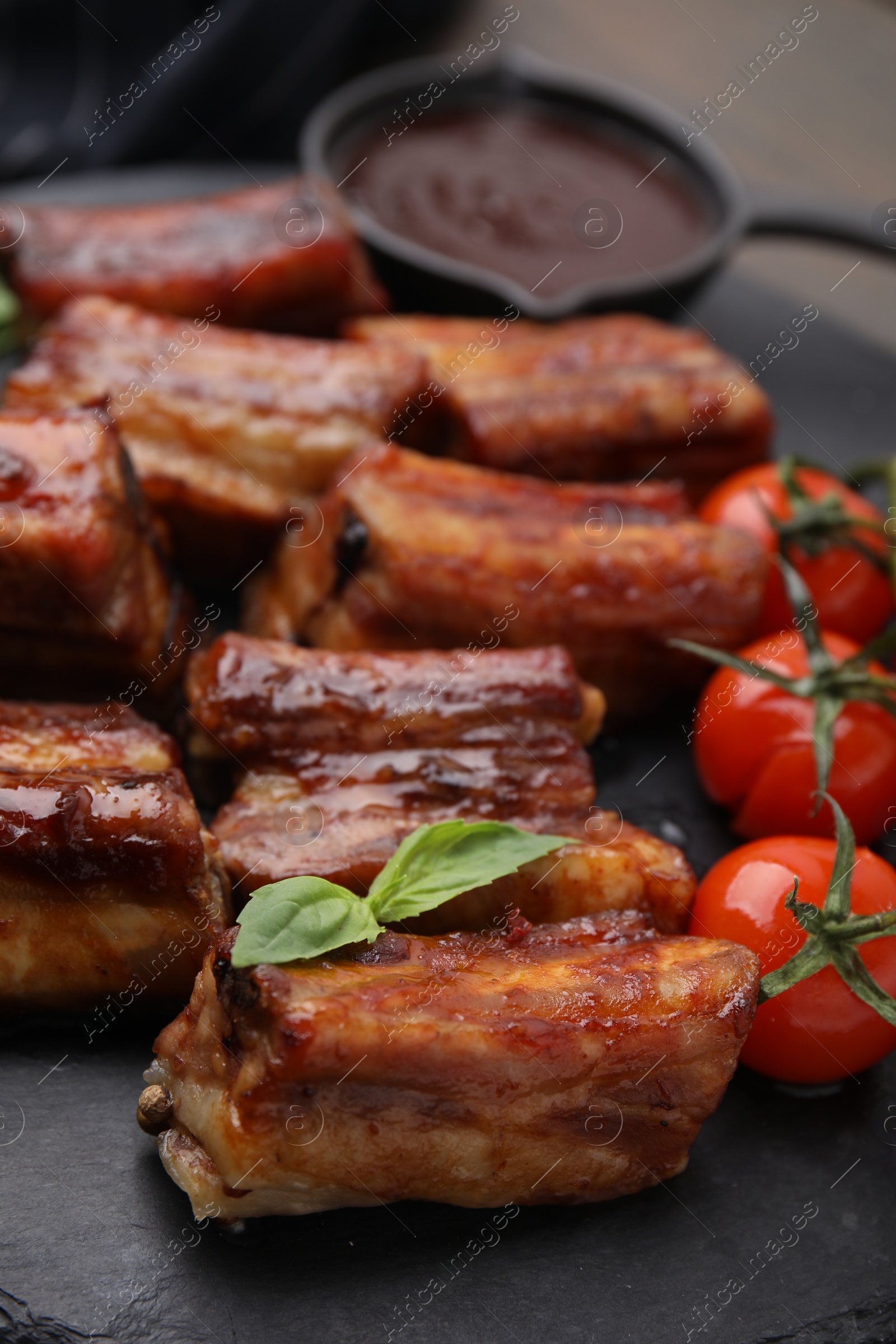 Photo of Tasty roasted pork ribs served with sauce, basil and tomatoes on table, closeup