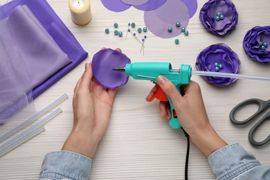 Photo of Woman with hot glue gun making textile flower at white wooden table, top view