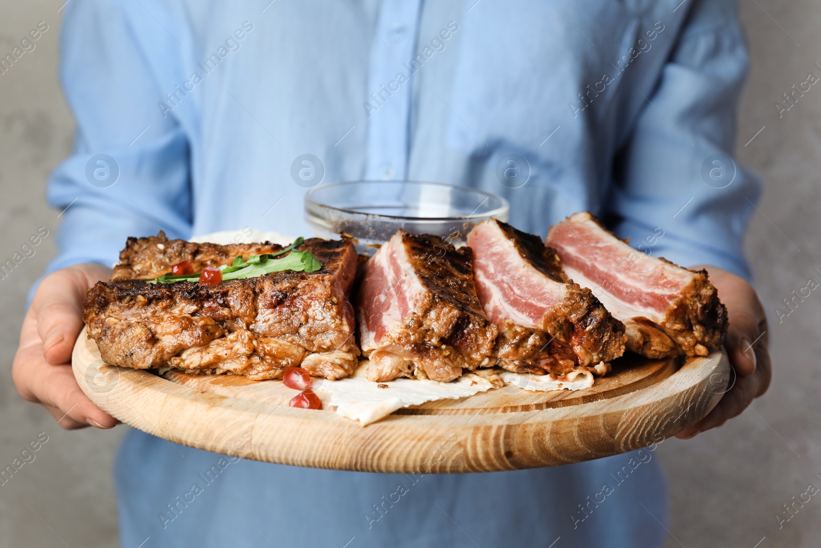 Photo of Woman holding wooden board with delicious grilled ribs on grey background, closeup