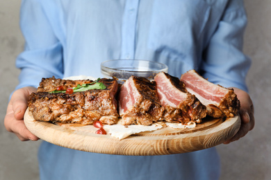 Photo of Woman holding wooden board with delicious grilled ribs on grey background, closeup