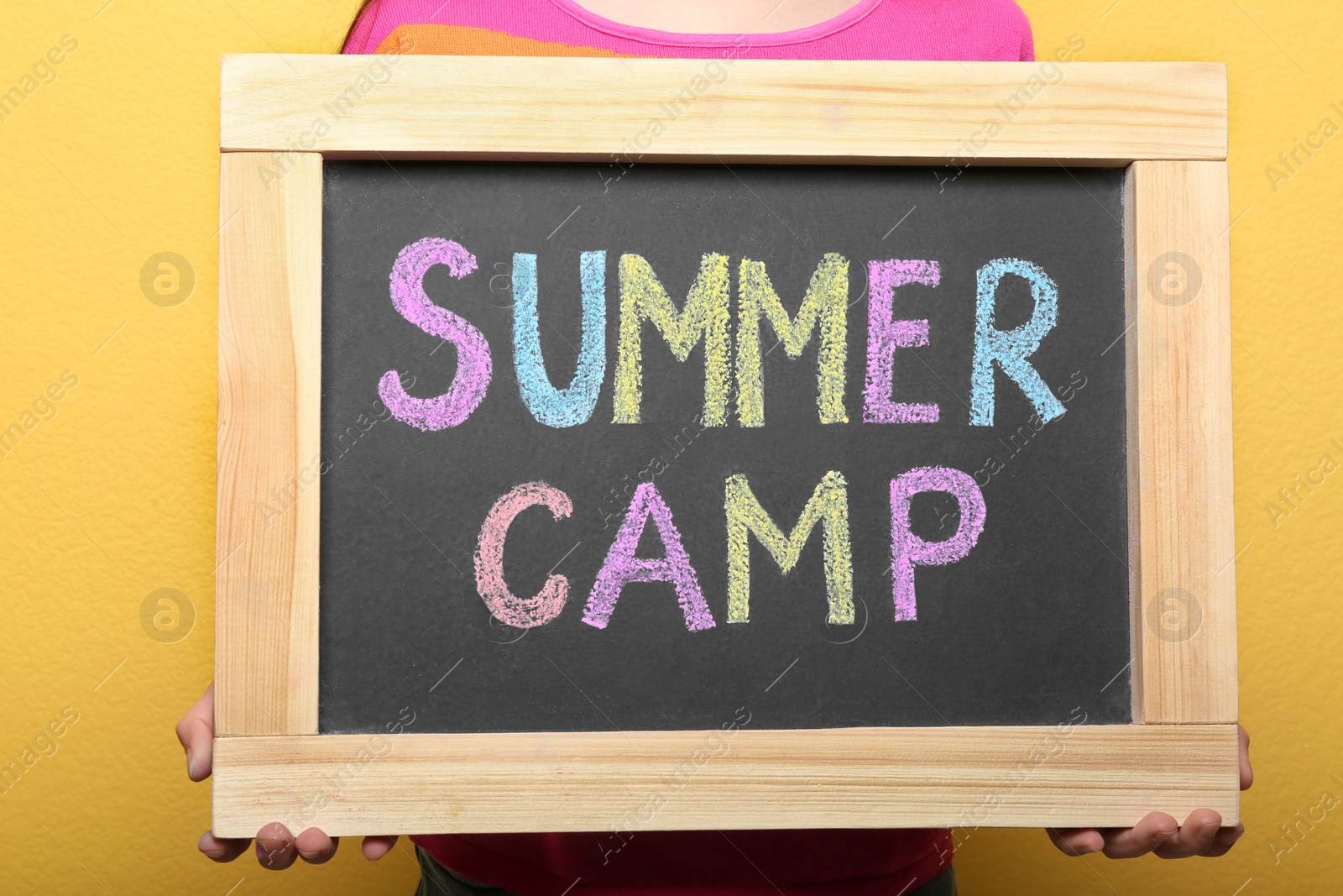 Photo of Child holding blackboard with words SUMMER CAMP chalked in different colors, closeup