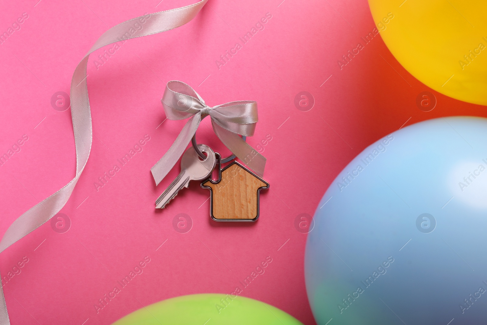 Photo of Key with trinket in shape of house and bow on pink background near color balloons, flat lay. Housewarming party