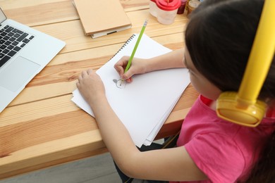 Photo of Little girl with headphones drawing on paper at online lesson indoors, above view. Distance learning