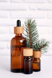 Photo of Bottles of essential oil and pine branch on white wooden table near brick wall