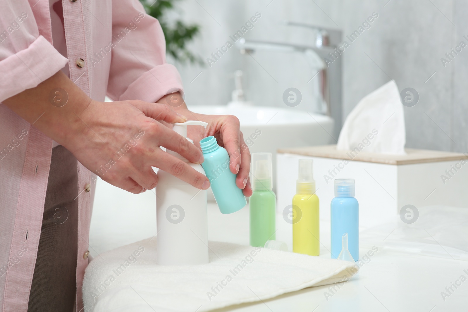 Photo of Woman pouring cosmetic product into plastic bottle at white countertop in bathroom, closeup. Bath accessories