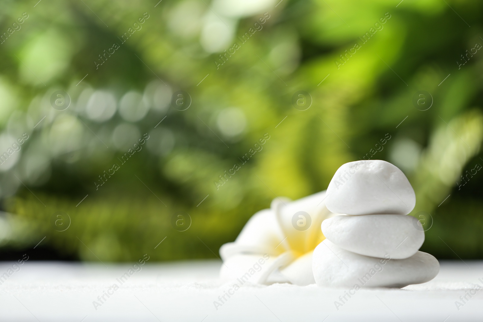 Photo of Stack of white stones and beautiful flower on sand against blurred green background, space for text. Zen, meditation, harmony