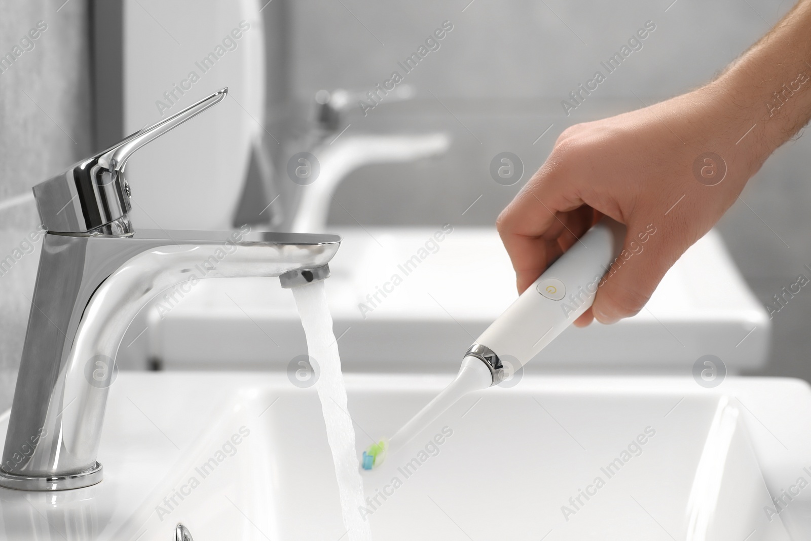 Photo of Man holding electric toothbrush near flowing water above sink in bathroom, closeup