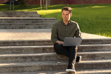 Handsome man with laptop sitting on concrete stairs outdoors. Space for text