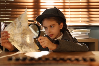 Photo of Cute little detective exploring document with magnifying glass at table in office