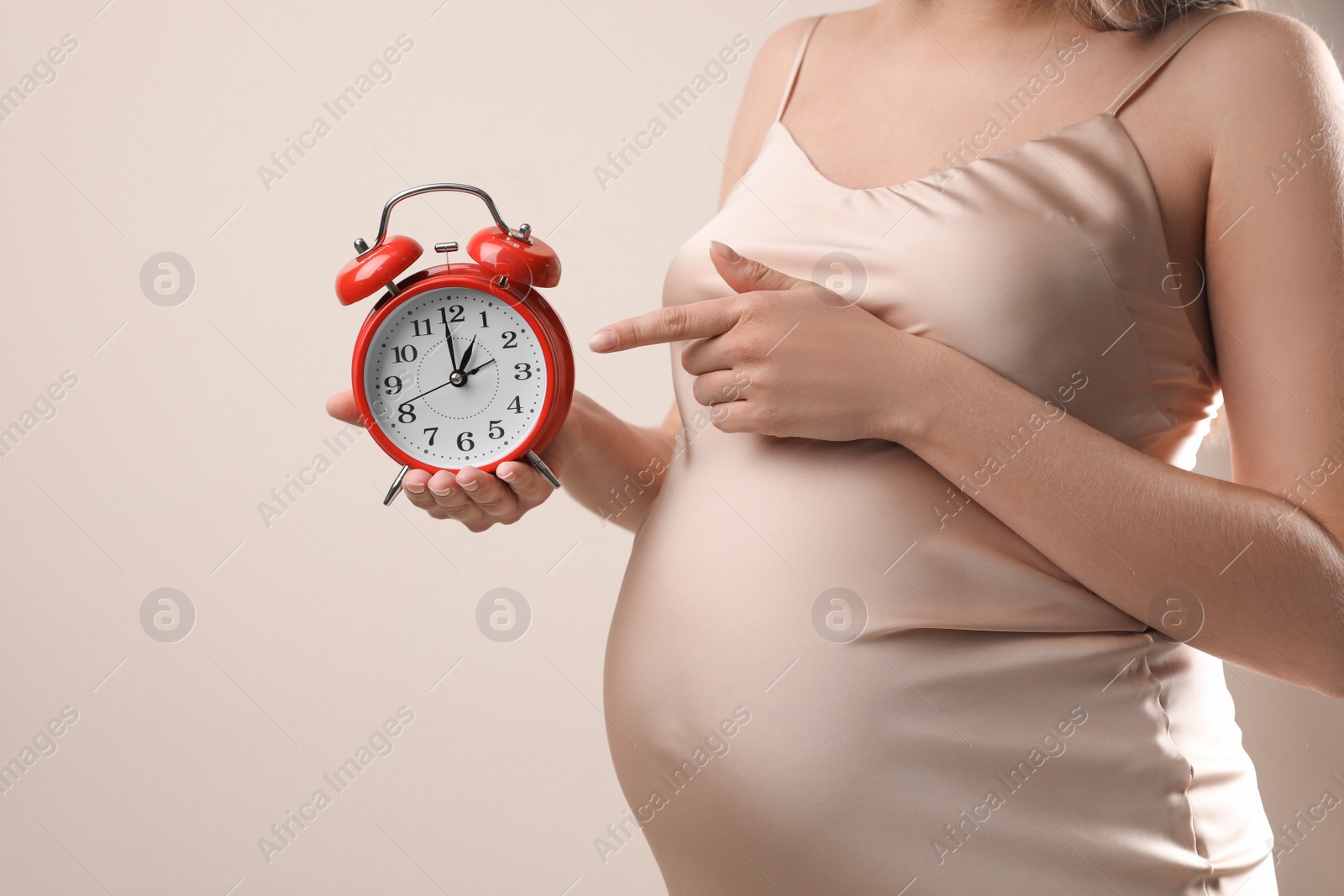 Photo of Young pregnant woman pointing at alarm clock near her belly on beige background, closeup. Time to give birth