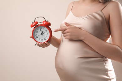 Young pregnant woman pointing at alarm clock near her belly on beige background, closeup. Time to give birth