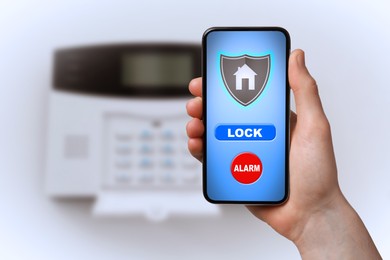 Woman operating home alarm system via mobile phone against white wall with security control panel, closeup. Application interface on device screen