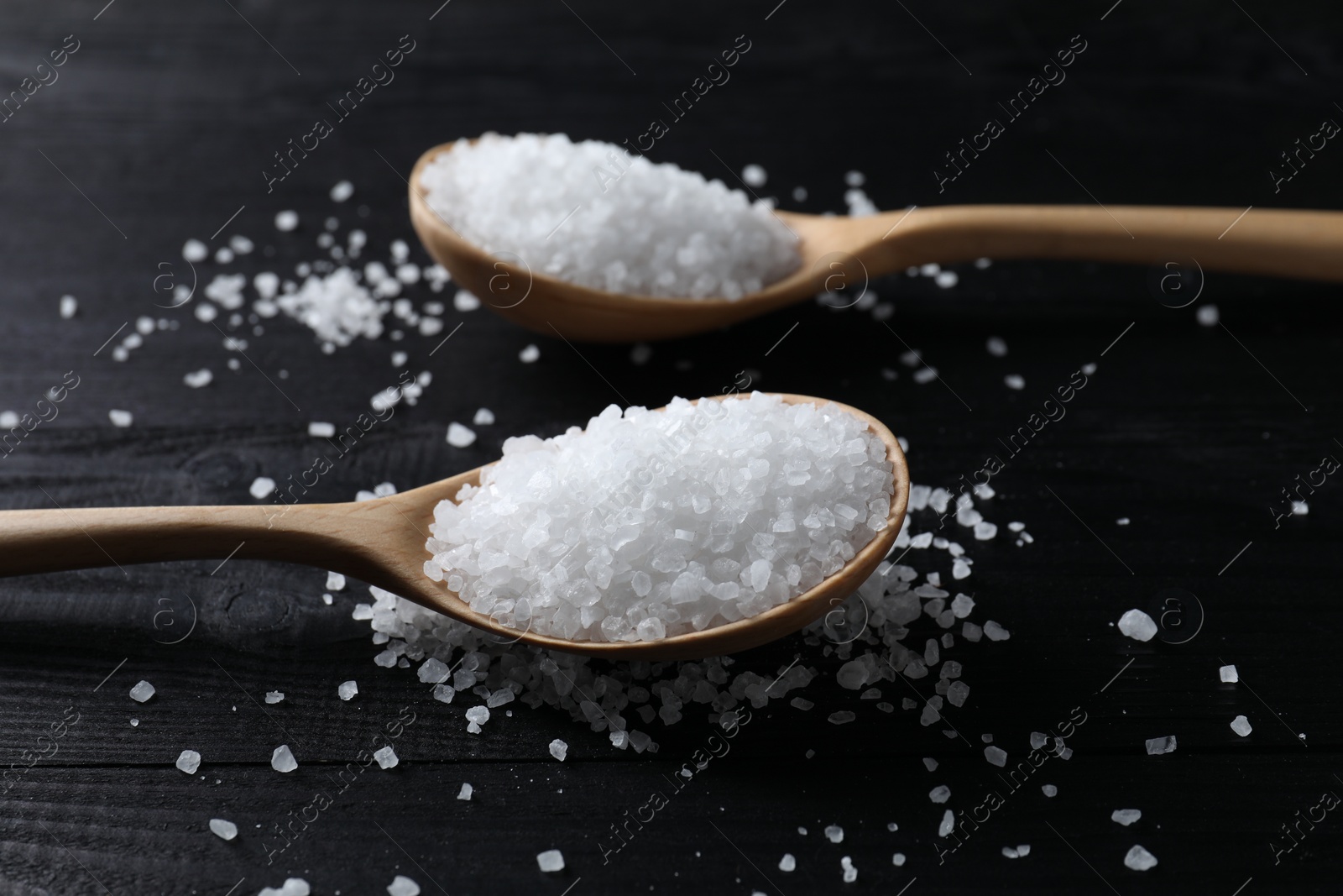 Photo of Organic salt in spoons on black wooden table, closeup