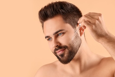 Handsome man applying cosmetic serum onto face on beige background, space for text