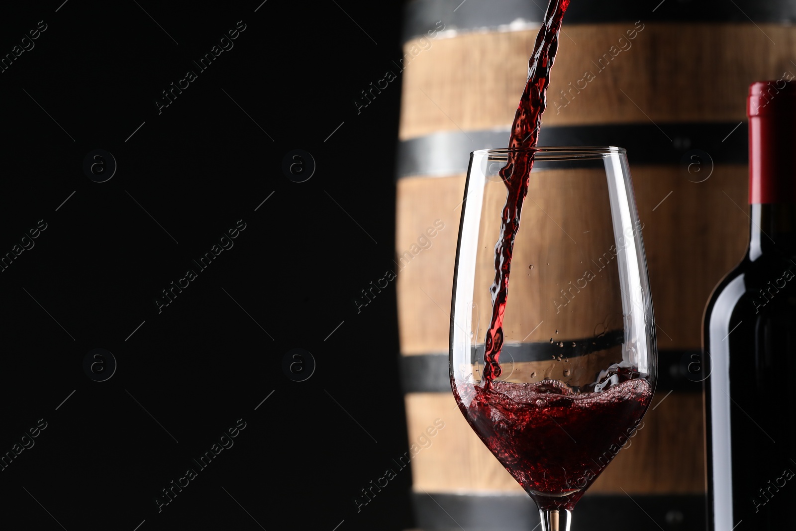 Photo of Pouring red wine into glass near wooden barrel against black background. Space for text