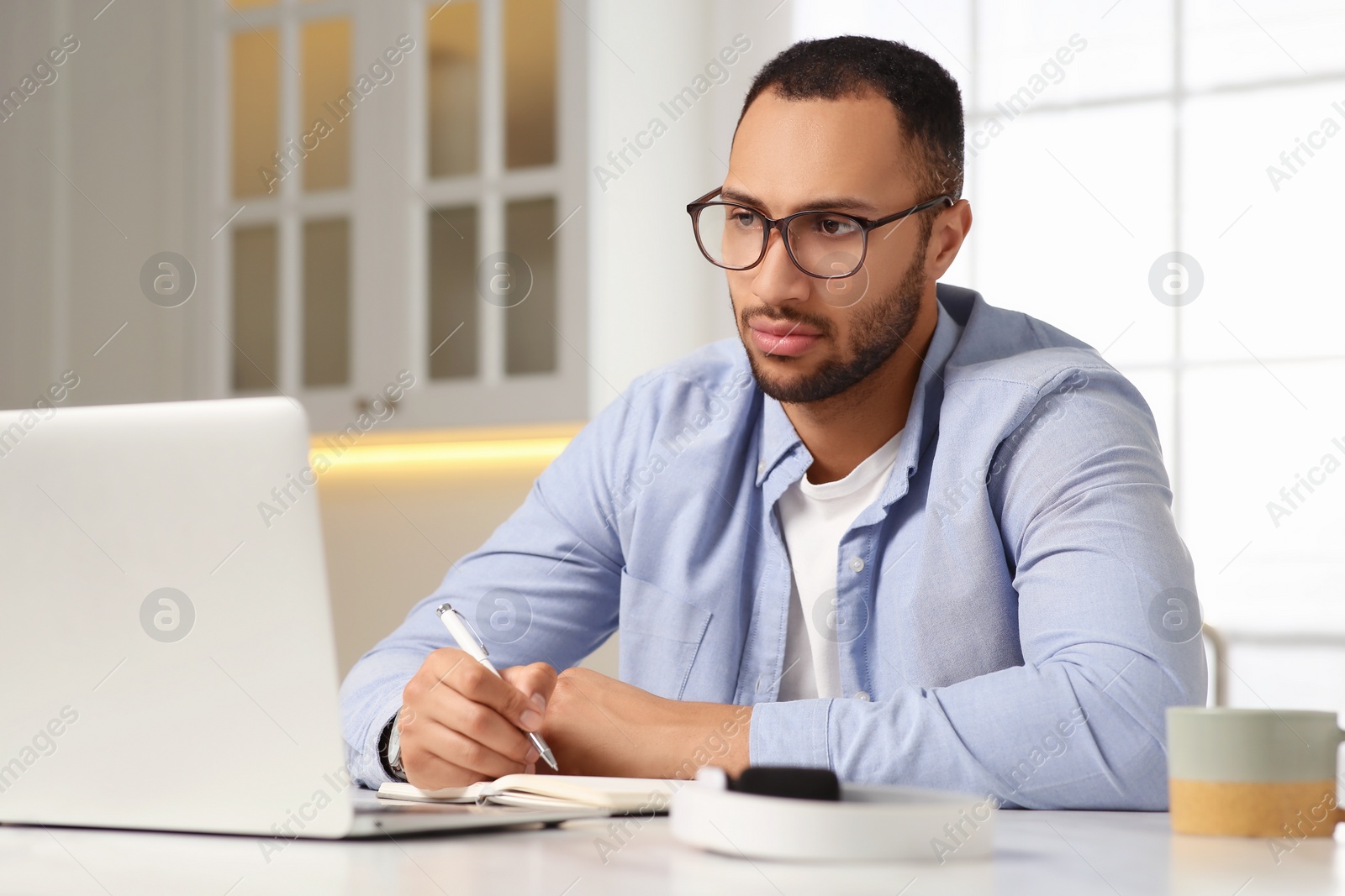 Photo of Young man with laptop writing in notebook at desk in kitchen. Home office