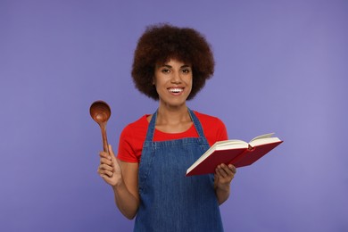 Photo of Happy young woman in apron holding spoon and recipe book on purple background