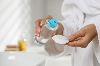 Photo of Woman pouring makeup remover from bottle onto cotton pad indoors, closeup