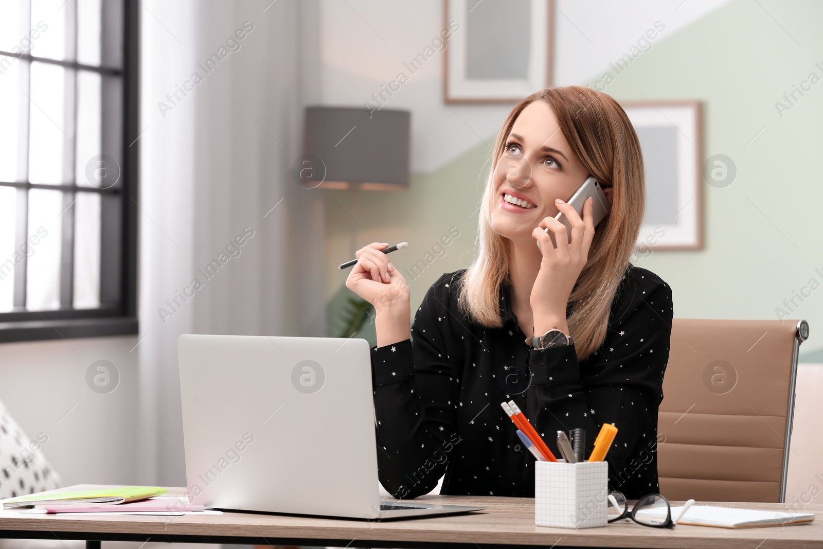 Photo of Young woman talking on phone while working with laptop at desk in home office