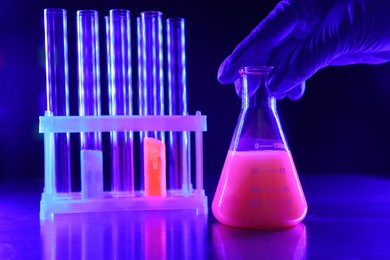 Photo of Scientist working with laboratory glassware of luminous liquids at table against dark background, closeup