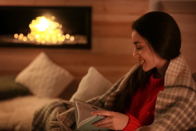 Photo of Woman reading book near decorative fireplace at home. Winter season