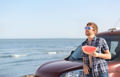 Young man with watermelon slice near car on beach. Space for text