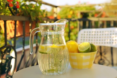 Jug with refreshing lemon water and citrus fruits in bowl on light table outdoors