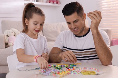 Photo of Happy father with his cute daughter making beaded jewelry at table in room