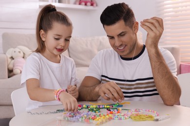 Happy father with his cute daughter making beaded jewelry at table in room