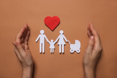 Photo of Woman protecting paper family figures and red heart on brown background, top view. Insurance concept