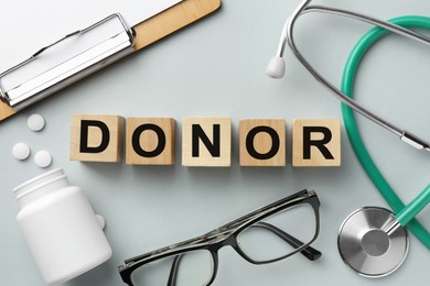 Photo of Word Donor made of wooden cubes, clipboard, stethoscope, glasses and pills on light grey table, flat lay