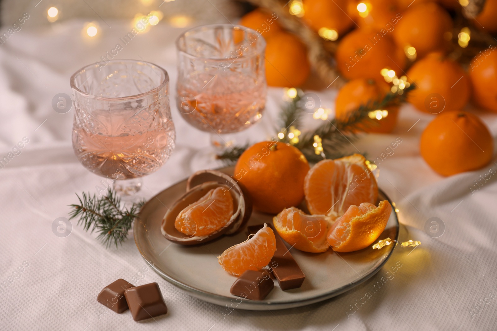 Photo of Delicious ripe tangerines, chocolates, festive lights and glasses of wine on white bedsheet