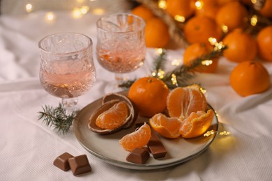 Delicious ripe tangerines, chocolates, festive lights and glasses of wine on white bedsheet