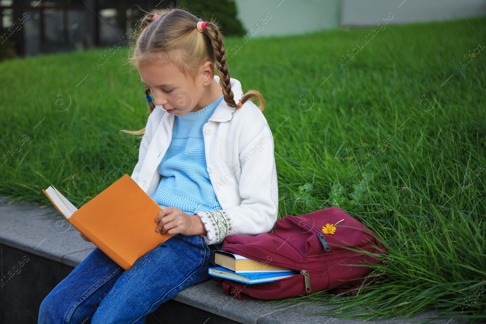 Photo of Cute little girl with backpack reading textbook on city street. Space for text