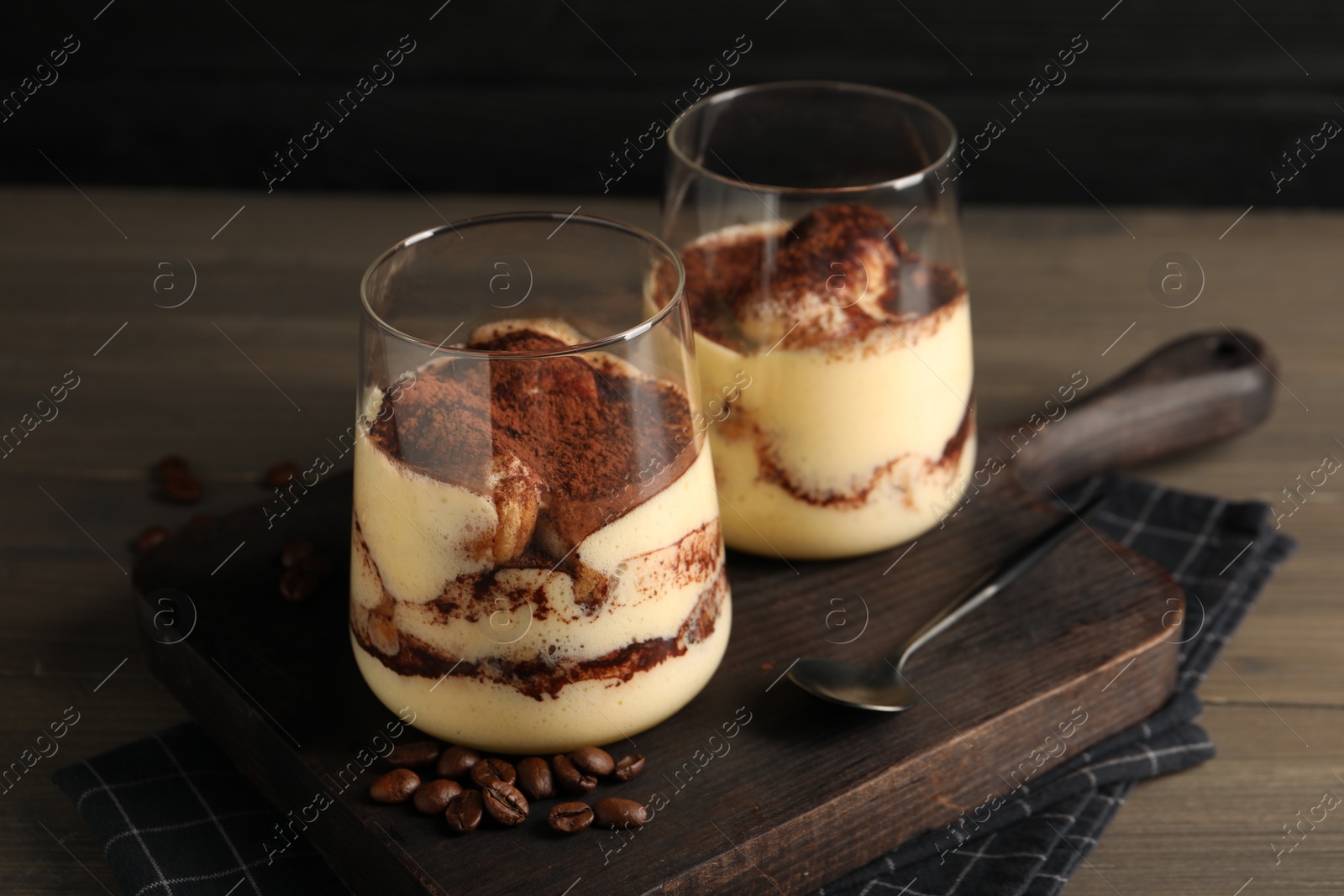 Photo of Delicious tiramisu in glasses, spoon and coffee beans on wooden table, closeup