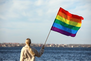 Photo of Woman holding bright LGBT flag near river, back view