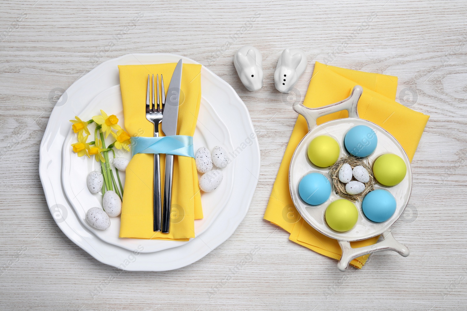 Photo of Festive table setting with painted eggs and cutlery on white wooden background, flat lay. Easter celebration