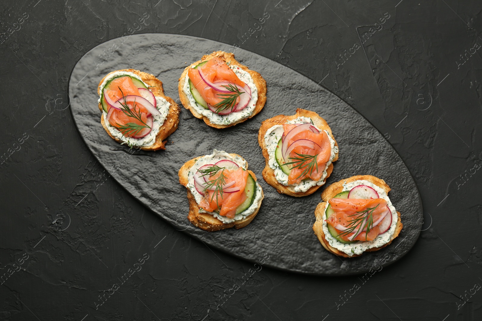 Photo of Tasty canapes with salmon, cucumber, radish and cream cheese on black table, top view