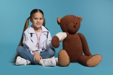 Photo of Little girl in medical uniform and toy bear with bandage on light blue background