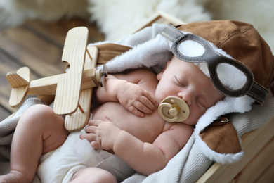 Cute newborn baby wearing aviator hat with toy sleeping in wooden crate