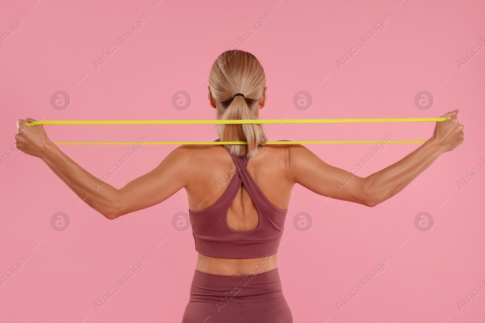 Photo of Woman exercising with elastic resistance band on pink background, back view