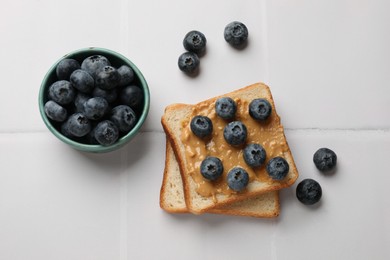 Photo of Delicious toasts with peanut butter and blueberries on white tiled table, flat lay