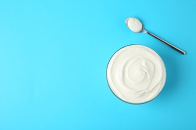 Photo of Glass bowl of sour cream and spoon on light blue background, flat lay. Space for text