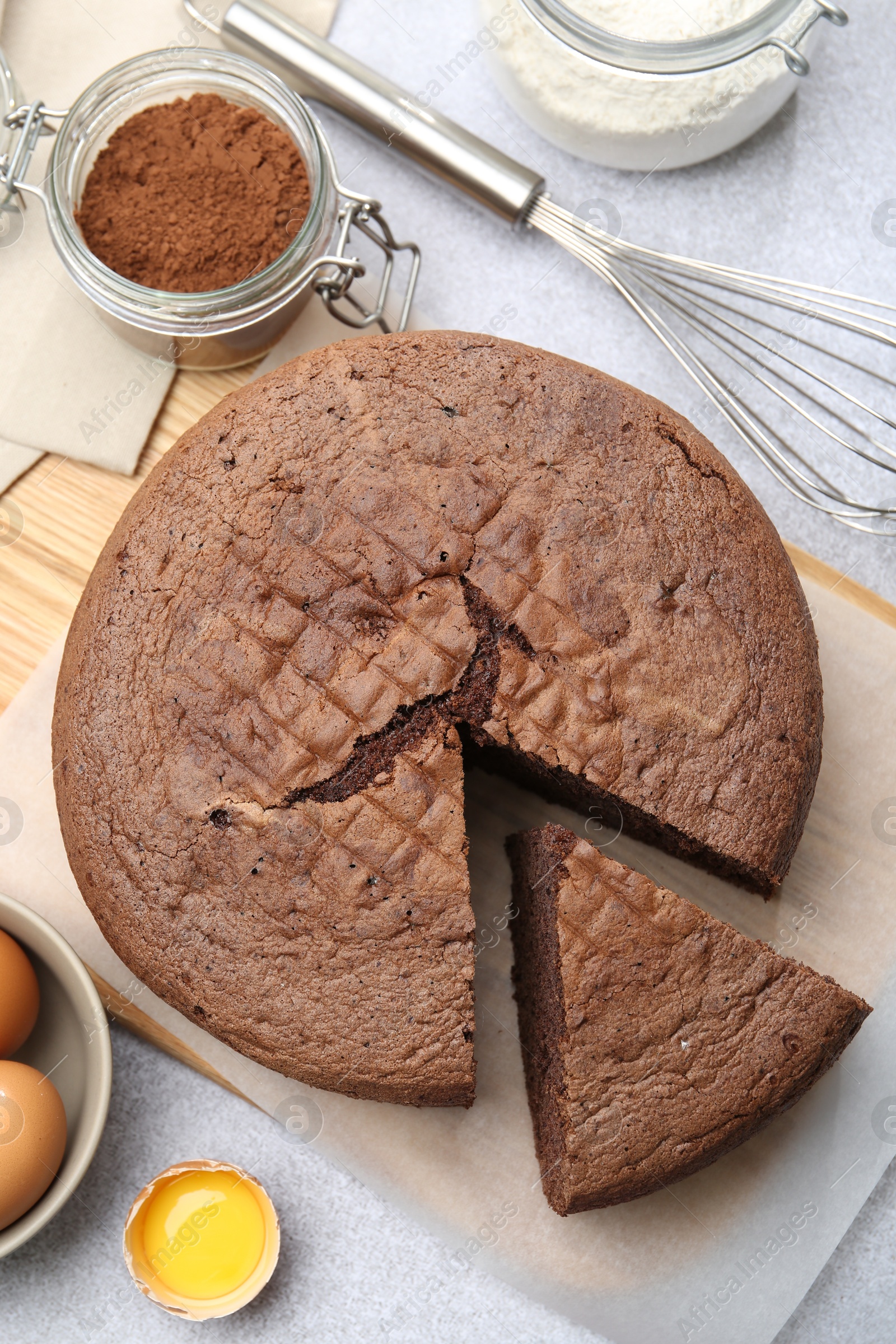 Photo of Cut chocolate sponge cake, whisk and ingredients on light table, flat lay