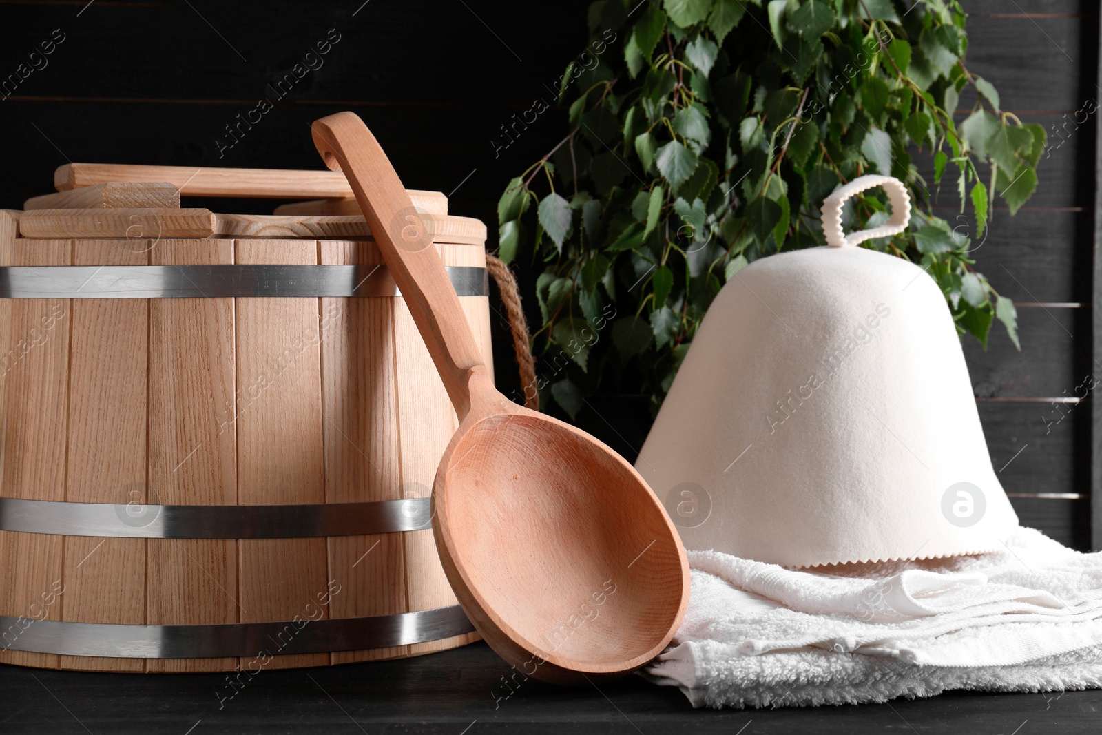 Photo of Sauna equipment. Wooden bucket, towel, felt wool hat and ladle on black surface