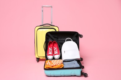 Photo of Two color suitcases with clothes, backpack and sneakers on pink background